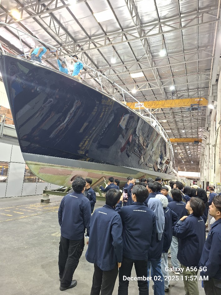 Students observing a large yacht hull under construction inside a shipyard hangar.